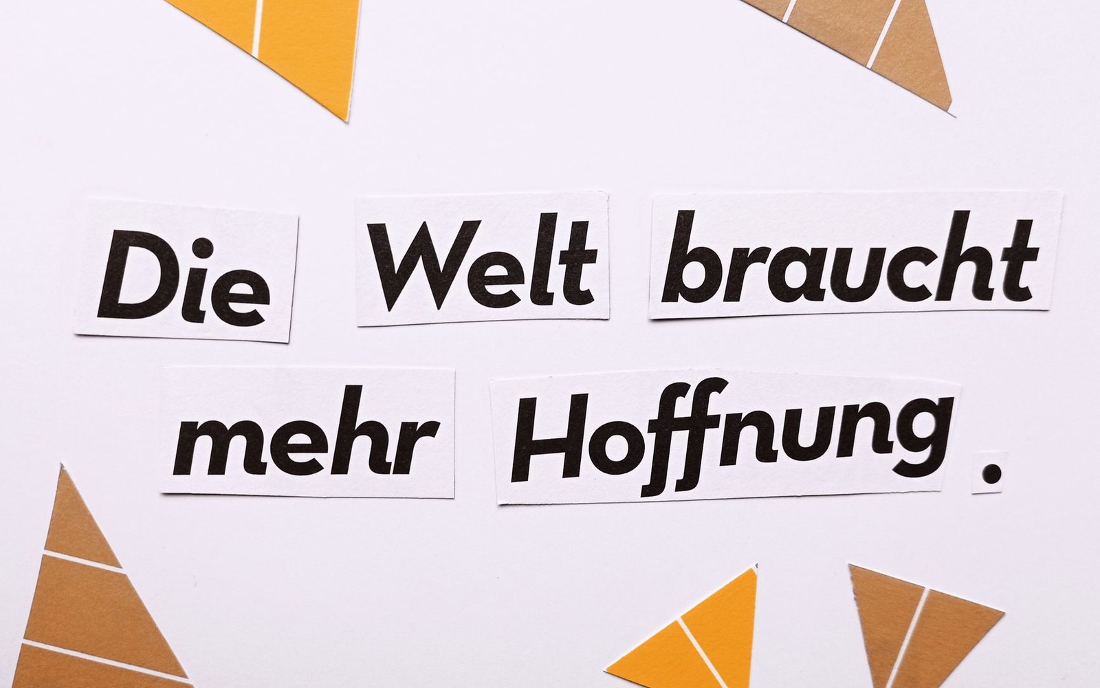 Cut-out paper letters spell "Die Welt braucht mehr Hoffnung" among yellow and brown triangles